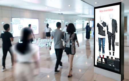 Görüntü tanıtımıyla ilgili detaylı açıklama örneği: Bir alışveriş merkezinde aynısı ve boyutları belirlenmiş yüz tanıma teknolojisi ile yaş ve cinsiyet tahmini yapan digital reklam ekranı yer almakta. Ekranın yanı sıra insanlar, cinsiyet ve yaş tahmini yapan yüz tanıma teknolojisini kullanmakta. Modern mağaza ve alışveriş ortamlarında yüz tanıma teknolojisinin kullanımı ve mağaza içi müşteri analitikleri üzerine odaklanan işlevsel bir görsel.