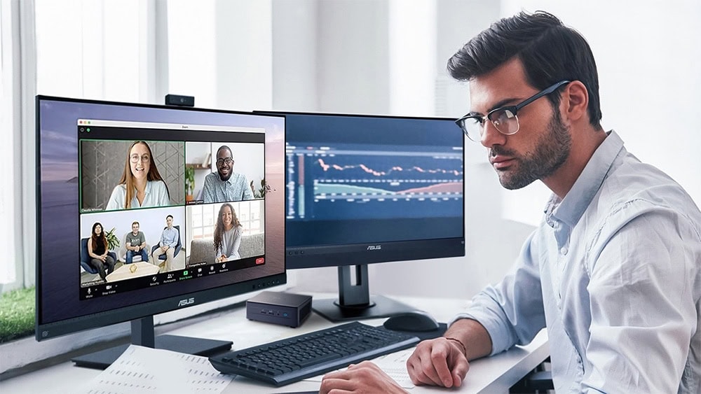 Çevrimiçi toplantıya katılan bozuk ve düzgün aydınlatılmış ofis ortamındaki genç adam, iki monitör, webcam, klavye ve not defteri ile çalışan profesyonel.