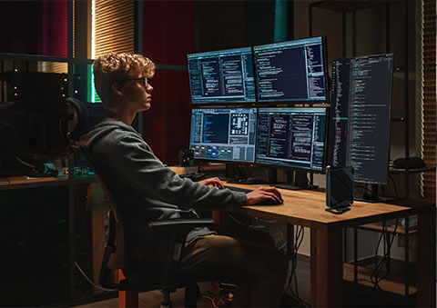 Yazılım geliştirme ve programlama yapan genç erkek, çok monitörlü bilgisayar ve kodlama ekranlarıyla oyun ve teknoloji odaklı çalışma ortamında.
